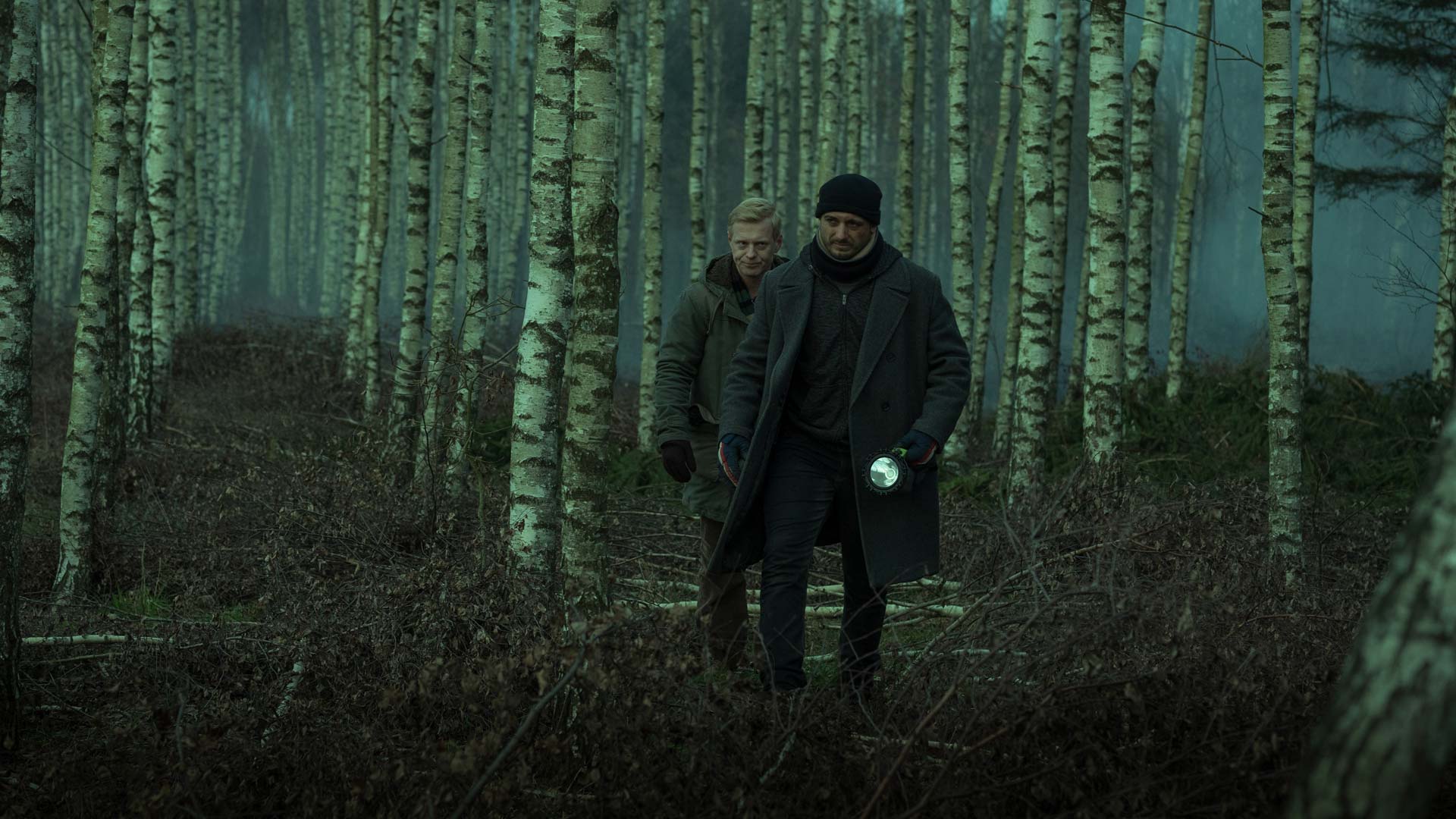 Auf dem Bild zu den internationalen Sendern und TV-Paketen bei Vodafone ist eine düstere Szene aus der polnischen Crime-Serie „Kruk“ zu sehen. Zwei Männer gehen durch einen nebligen Birkenwald, einer hält eine Taschenlampe in der Hand.