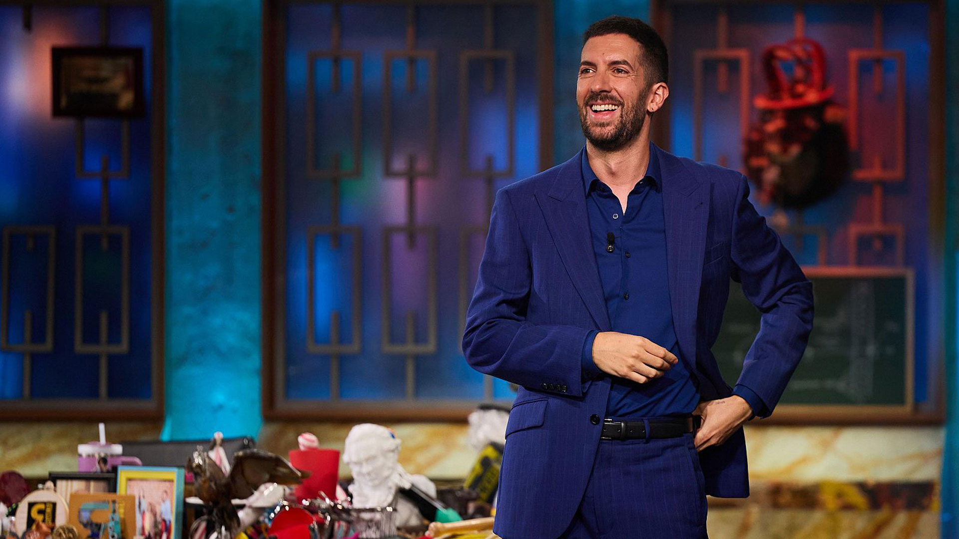 Auf dem Bild zu den internationalen Sendern und TV-Paketen bei Vodafone ist Moderator David Broncano aus der spanischen Comedy-Show „La Revuelta“ zu sehen. Er trägt einen blauen Anzug und steht lachend in einem bunt beleuchteten Studio mit dekorativem Hintergrund.