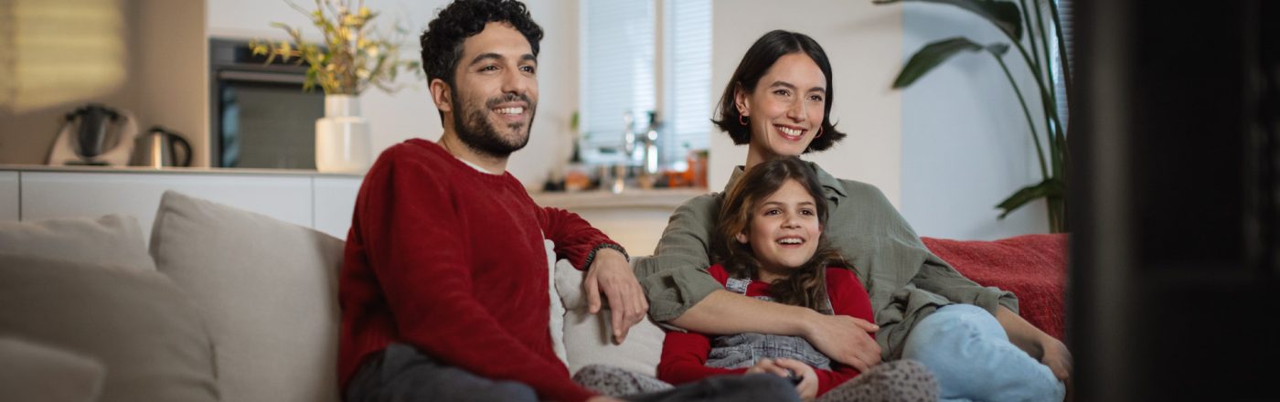 Auf dem Bild zu den internationalen Sendern und TV-Paketen bei Vodafone sitzt eine fröhliche Familie gemeinsam auf dem Sofa und schaut entspannt Fernsehen. Ein Mann, eine Frau und ein Mädchen lachen und wirken völlig in das TV-Programm vertieft.