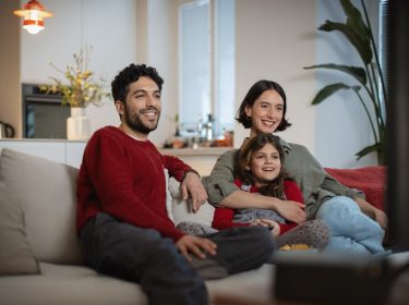Auf dem Bild zu den internationalen Sendern und TV-Paketen bei Vodafone sitzt eine fröhliche Familie gemeinsam auf dem Sofa und schaut entspannt Fernsehen. Ein Mann, eine Frau und ein Mädchen lachen und wirken völlig in das TV-Programm vertieft.
