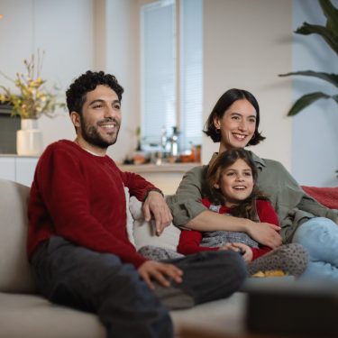 Auf dem Bild zu den internationalen Sendern und TV-Paketen bei Vodafone sitzt eine fröhliche Familie gemeinsam auf dem Sofa und schaut entspannt Fernsehen. Ein Mann, eine Frau und ein Mädchen lachen und wirken völlig in das TV-Programm vertieft.