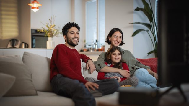 Auf dem Bild zu den internationalen Sendern und TV-Paketen bei Vodafone sitzt eine fröhliche Familie gemeinsam auf dem Sofa und schaut entspannt Fernsehen. Ein Mann, eine Frau und ein Mädchen lachen und wirken völlig in das TV-Programm vertieft.