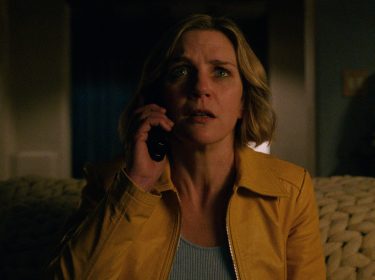 Auf dem Bild aus der Serie Pluribus ist Rhea Seehorn in ihrer Rolle zu sehen. Sie trägt eine gelbe Jacke und hält ein Telefon ans Ohr, während sie mit besorgtem Blick in die Ferne schaut. Die Beleuchtung ist düster.