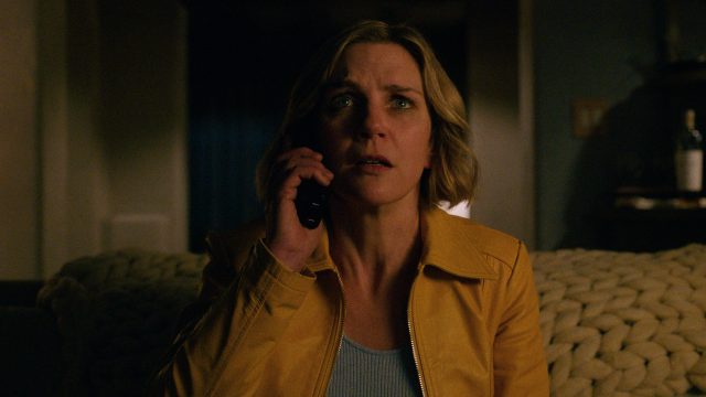 Auf dem Bild aus der Serie Pluribus ist Rhea Seehorn in ihrer Rolle zu sehen. Sie trägt eine gelbe Jacke und hält ein Telefon ans Ohr, während sie mit besorgtem Blick in die Ferne schaut. Die Beleuchtung ist düster.