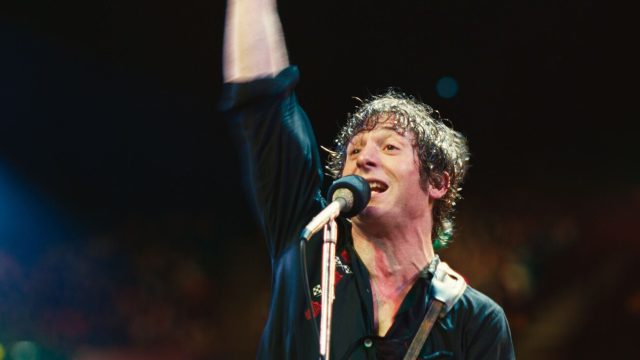 Jeremy Allen White wird in „Springsteen: Deliver Me From Nowhere”: Ein durchgeschwitzter Mann steht mit erhobenem Arm und Gitarrengurt über der Schulter auf einer Bühne vor einem Mikrofon.