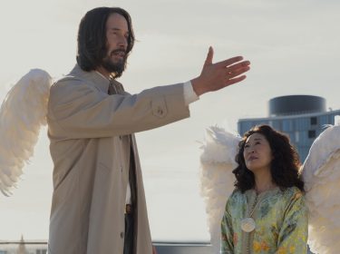 Keanu Reeves in Good Fortune: Ein Mann mit weißen Engelsflügeln und beigem Trenchcoat, gestikuliert gegenüber einer gelangweilten Frau, die ebenfalls Engelsflügel trägt.