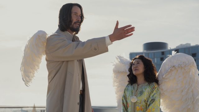 Keanu Reeves in Good Fortune: Ein Mann mit weißen Engelsflügeln und beigem Trenchcoat, gestikuliert gegenüber einer gelangweilten Frau, die ebenfalls Engelsflügel trägt.