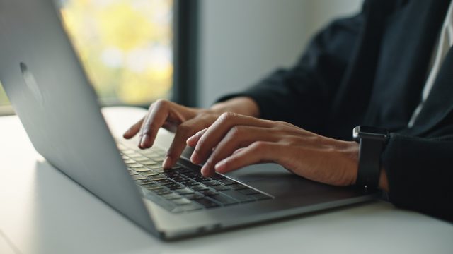 Auf dem Bild mit symbolischem Bezug zu Perplexity Pro ist eine Person in Business-Kleidung zu sehen, die konzentriert auf einem modernen Laptop tippt. Die Szene wirkt professionell und fokussiert, im Hintergrund ist ein Fenster mit natürlichem Licht und unscharfem Ausblick zu erkennen. Am Handgelenk trägt die Person eine Smartwatch.