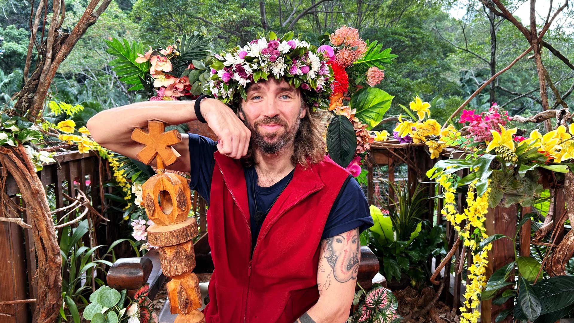 Auf dem Bild zum Dschungelcamp Gewinner 2026 ist Gil Ofarim zu sehen, der entspannt und zufrieden in einer üppigen Dschungelkulisse sitzt. Er trägt eine rote Weste, ein Blumenkranz schmückt sein Haar, in der Hand hält er das Sieger-Totem. Die Stimmung wirkt triumphierend, naturverbunden und feierlich.