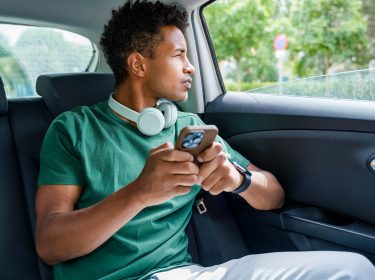 Ein junger Mann sitzt in einem Auto auf einer Rücksitzbank, schaut aus dem Fenster und hält ein Smartphone in der Hand.