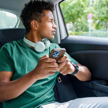 Ein junger Mann sitzt in einem Auto auf einer Rücksitzbank, schaut aus dem Fenster und hält ein Smartphone in der Hand.