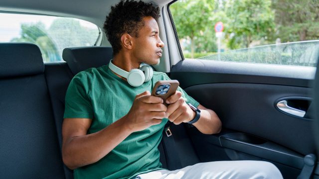 Ein junger Mann sitzt in einem Auto auf einer Rücksitzbank, schaut aus dem Fenster und hält ein Smartphone in der Hand.
