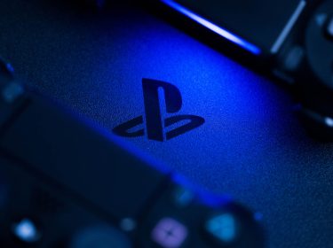 Nahaufnahme des PlayStation-Logos auf einer Konsole, beleuchtet von blauem Licht, mit zwei teilweise sichtbaren Controllern im Vorder- und Hintergrund.