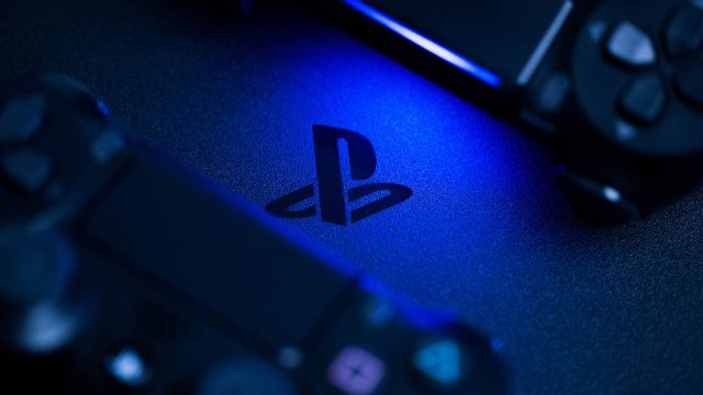 Nahaufnahme des PlayStation-Logos auf einer Konsole, beleuchtet von blauem Licht, mit zwei teilweise sichtbaren Controllern im Vorder- und Hintergrund.