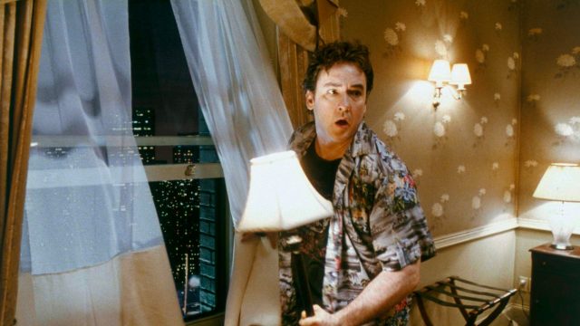 Filmszene aus Zimmer 1408: Mike Enslin (John Cusack) steht mit einer Lampe in der Hand vor dem geöffneten Fenster und schaut sich ängstlich um.
