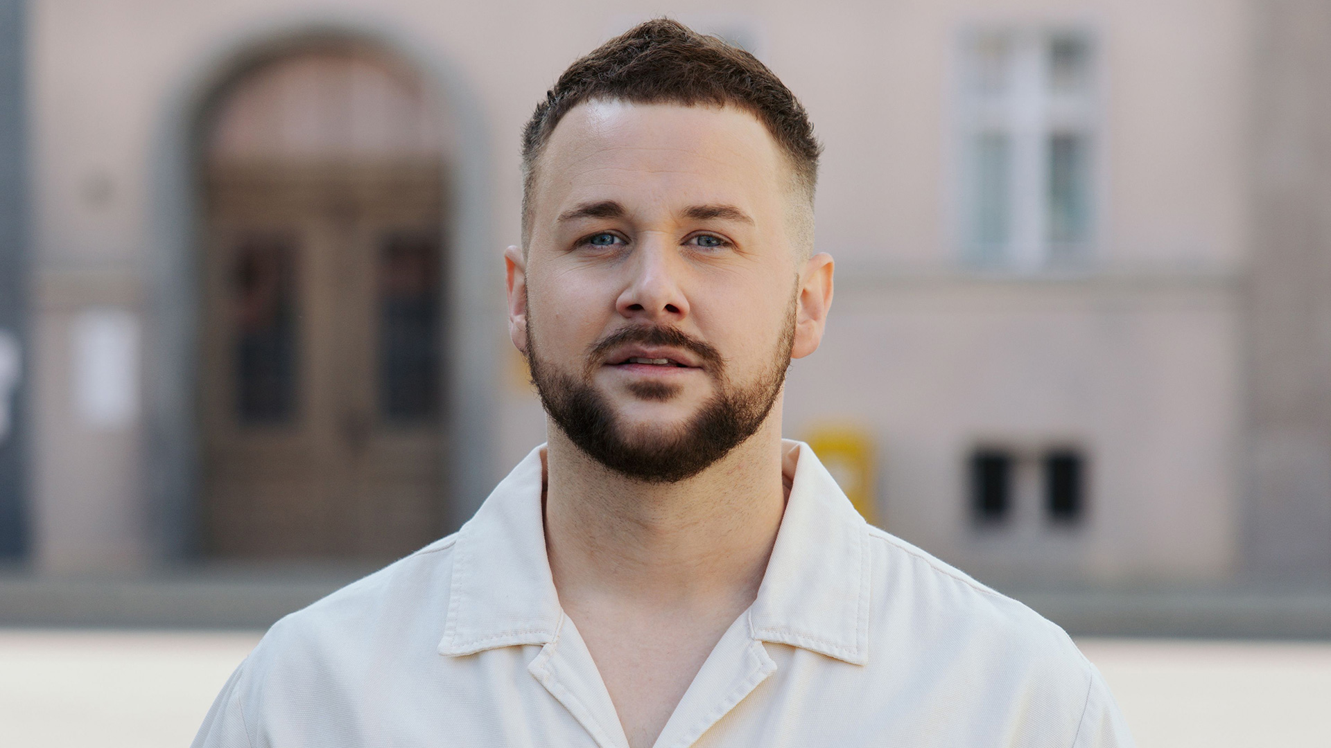 Das Bild zur GZSZ-Besetzung zeigt Felix von Deventer. Er steht im Freien vor einem Gebäude und trägt ein helles Hemd.