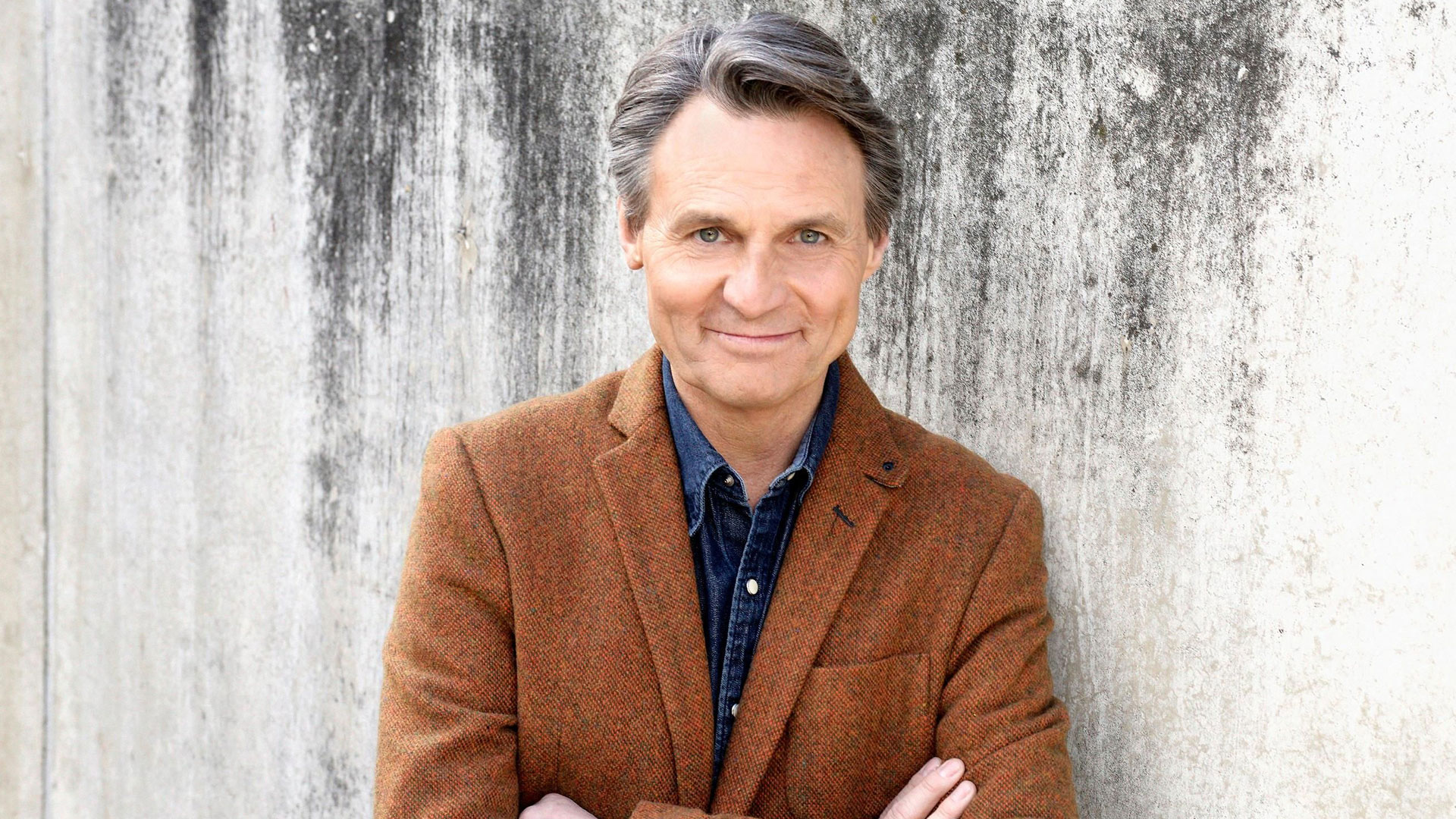 Dieses Bild zur GZSZ-Besetzung zeigt Wolfgang Bahro. Er steht mit verschränkten Armen vor einer strukturierten Wand, trägt ein braunes Sakko über einem Jeanshemd und lächelt in die Kamera.