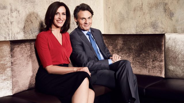 Auf dem Bild zur GZSZ-Besetzung sitzen Ulrike Frank und Wolfgang Bahro nebeneinander auf einem Sofa. Ulrike Frank trägt eine rote Bluse, Wolfgang Bahro einen grauen Anzug.