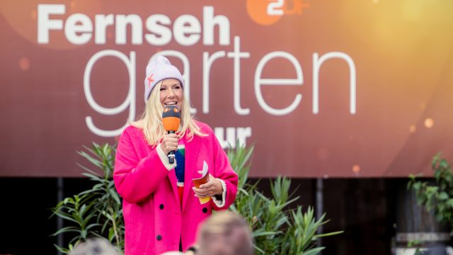 "ZDF-Fernsehgarten on tour": Andrea Kiewel steht in einem pinkfarbenen Mantel vor dem Sendungslogo. Sie hält ein Mikrofon und Moderationskarten in der Hand. Sie schaut lächelnd in Richtung Publikum.