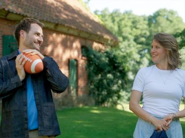 Auf dem Bild zur Besetzung von Rote Rosen in Staffel 24 sind Daniel, gespielt von Daniel Fritz, und Lou, dargestellt von Eva-Maria Grein von Friedl, in einer sommerlichen Szene im Grünen zu sehen. Beide blicken sich lächelnd an, während Daniel einen Football hält. Die Stimmung wirkt leicht und verspielt, mit einem ländlichen Gebäude im Hintergrund.
