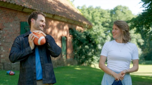 Auf dem Bild zur Besetzung von Rote Rosen in Staffel 24 sind Daniel, gespielt von Daniel Fritz, und Lou, dargestellt von Eva-Maria Grein von Friedl, in einer sommerlichen Szene im Grünen zu sehen. Beide blicken sich lächelnd an, während Daniel einen Football hält. Die Stimmung wirkt leicht und verspielt, mit einem ländlichen Gebäude im Hintergrund.