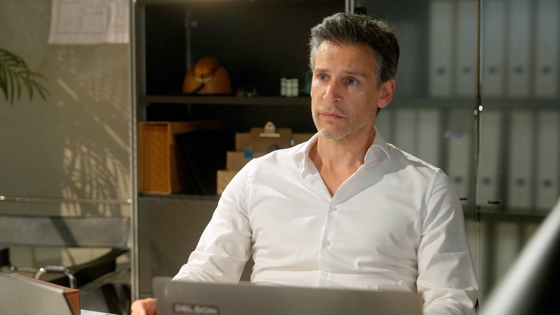 Im Bild zur Besetzung von Rote Rosen in Staffel 24 ist Richard, gespielt von Florian Odendahl, in einem modernen Büro zu sehen. Er trägt ein weißes Hemd und sitzt nachdenklich vor seinem Laptop. Die Szene wirkt ruhig und konzentriert, mit Aktenordnern und Regalen im Hintergrund, was auf ein berufliches Umfeld hinweist.