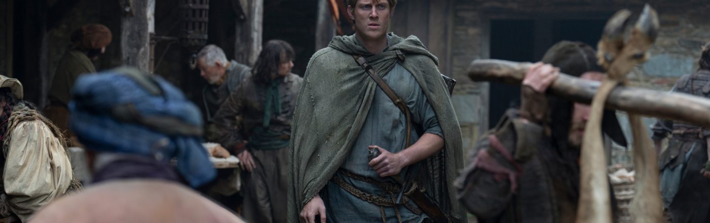 In dieser Szene aus A Knight of the Seven Kingdoms ist Heckenritter Dunk, gespielt von Peter Claffey, in einem mittelalterlichen Dorf zu sehen. Er trägt einen grünen Umhang und hält eine kleine Flasche in der Hand, während er mit entschlossenem Blick durch eine belebte Marktstraße geht. Die Umgebung wirkt rau und lebendig, mit einfachen Holzhäusern und geschäftigen Dorfbewohnern.