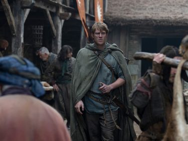 In dieser Szene aus A Knight of the Seven Kingdoms ist Heckenritter Dunk, gespielt von Peter Claffey, in einem mittelalterlichen Dorf zu sehen. Er trägt einen grünen Umhang und hält eine kleine Flasche in der Hand, während er mit entschlossenem Blick durch eine belebte Marktstraße geht. Die Umgebung wirkt rau und lebendig, mit einfachen Holzhäusern und geschäftigen Dorfbewohnern.