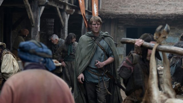 In dieser Szene aus A Knight of the Seven Kingdoms ist Heckenritter Dunk, gespielt von Peter Claffey, in einem mittelalterlichen Dorf zu sehen. Er trägt einen grünen Umhang und hält eine kleine Flasche in der Hand, während er mit entschlossenem Blick durch eine belebte Marktstraße geht. Die Umgebung wirkt rau und lebendig, mit einfachen Holzhäusern und geschäftigen Dorfbewohnern.