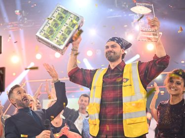 Auf dem Bild ist Jimi Blue Ochsenknecht als frisch gekürter Promi Big Brother Gewinner 2025 zu sehen. Er trägt eine gelbe Warnweste und hält stolz einen durchsichtigen Pokal sowie einen Koffer voller Geldscheine in die Höhe. Um ihn herum jubelt das Publikum, Konfetti fliegt durch die Luft, die Stimmung ist ausgelassen und feierlich.