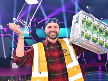 Der Sieger von „Promi Big Brother“ 2025 trägt ein kariertes Hemd, eine gelbe Weste und hält die Siegertrophäe sowie einen Geldkoffer hoch.