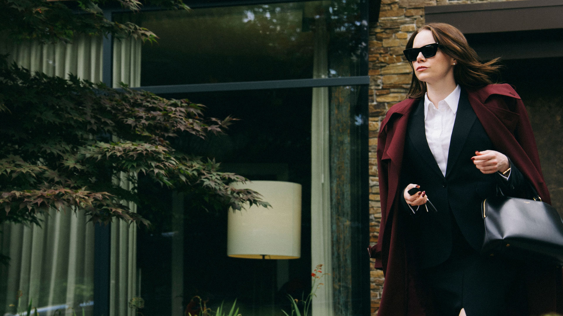 Emma Stone in Bugonia: Eine Frau in einem dunklen Business-Outfit und Sonnenbrille verlässt ein modernes Gebäude, eine Tasche und Autoschlüssel in der Hand.