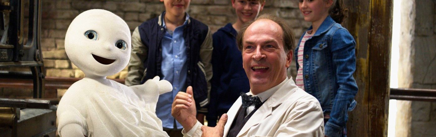 Auf dem Bild aus dem Film Das kleine Gespenst, einem Klassiker unter den Halloween-Filmen für die ganze Familie, ist das fröhlich dreinblickende Gespenst mit einem großen Schlüsselbund in der Luft schwebend zu sehen. Daneben steht ein lachender Mann in weißem Kittel, umgeben von drei neugierigen Kindern vor einer rustikalen Ziegelwand. Die Stimmung ist heiter und kindgerecht.