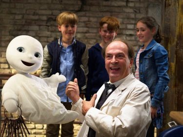 Auf dem Bild aus dem Film Das kleine Gespenst, einem Klassiker unter den Halloween-Filmen für die ganze Familie, ist das fröhlich dreinblickende Gespenst mit einem großen Schlüsselbund in der Luft schwebend zu sehen. Daneben steht ein lachender Mann in weißem Kittel, umgeben von drei neugierigen Kindern vor einer rustikalen Ziegelwand. Die Stimmung ist heiter und kindgerecht.