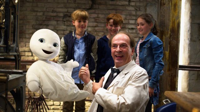 Auf dem Bild aus dem Film Das kleine Gespenst, einem Klassiker unter den Halloween-Filmen für die ganze Familie, ist das fröhlich dreinblickende Gespenst mit einem großen Schlüsselbund in der Luft schwebend zu sehen. Daneben steht ein lachender Mann in weißem Kittel, umgeben von drei neugierigen Kindern vor einer rustikalen Ziegelwand. Die Stimmung ist heiter und kindgerecht.