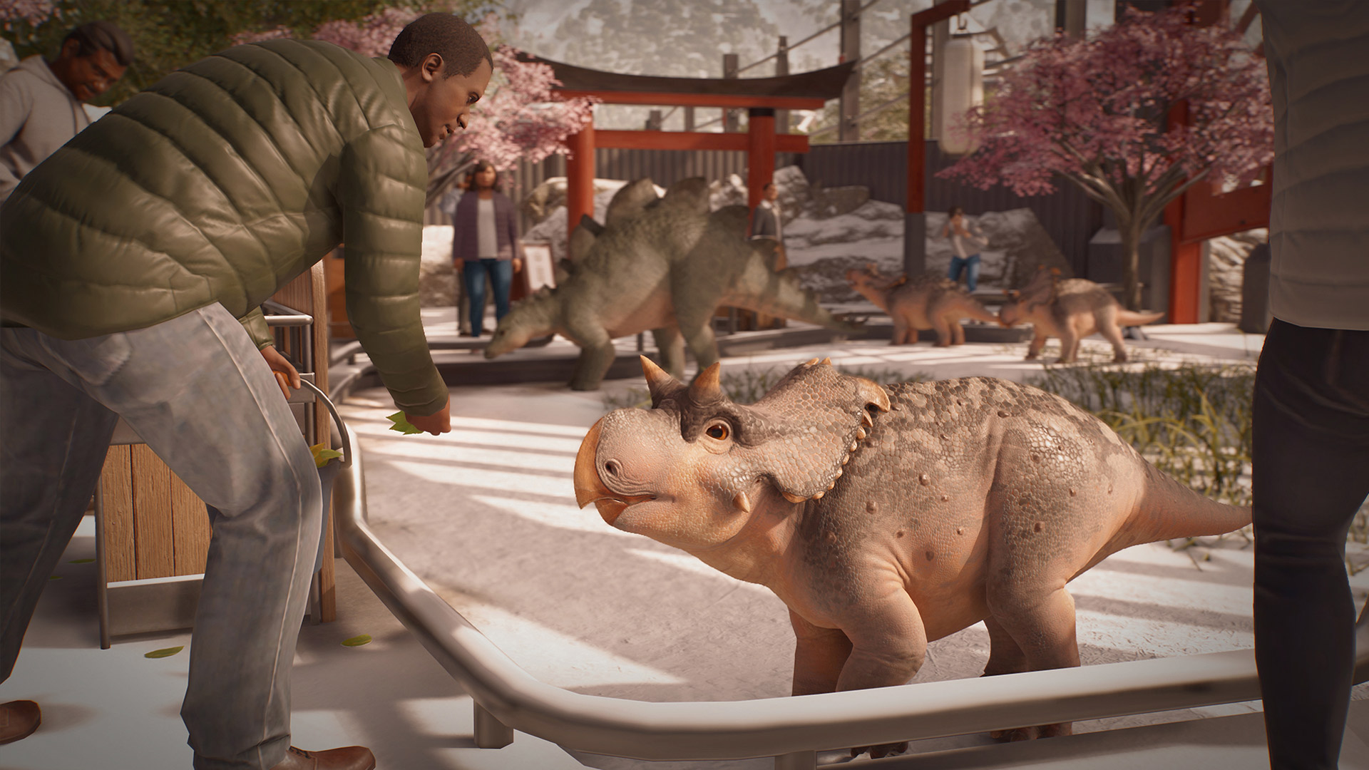 In Jurassic World Evolution 3 füttert ein Besucher ein junges Dinosaurierbaby durch ein Geländer, während im Hintergrund weitere kleine und große Dinosaurier durch ein Gehege mit Kirschbäumen laufen. Diese Szene zeigt eindrucksvoll die emotionale Tiefe und Interaktivität, die Jurassic World Evolution 3-Tipps für ein erfolgreiches Parkmanagement empfehlen.