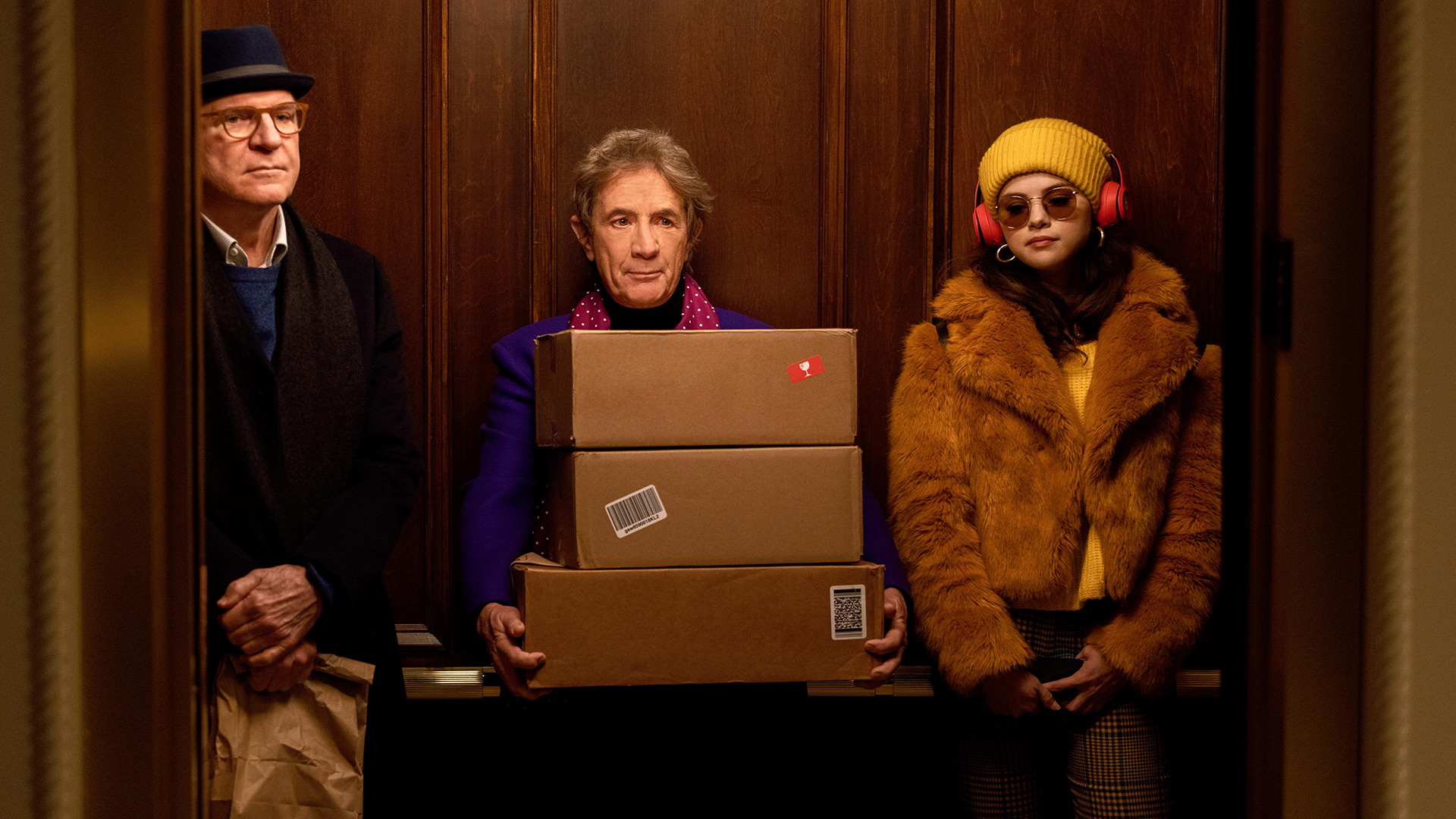 Auf dem Bild in dem Artikel zu Only Murders in the Building Staffel 6 stehen Charles, Oliver und Mabel in einem Aufzug. Charles trägt Hut und Mantel, Oliver hält drei Pakete im Arm und trägt ein violettes Sakko, Mabel steht in einer gelben Felljacke mit roter Kopfhörer-Mütze-Kombi und Sonnenbrille.