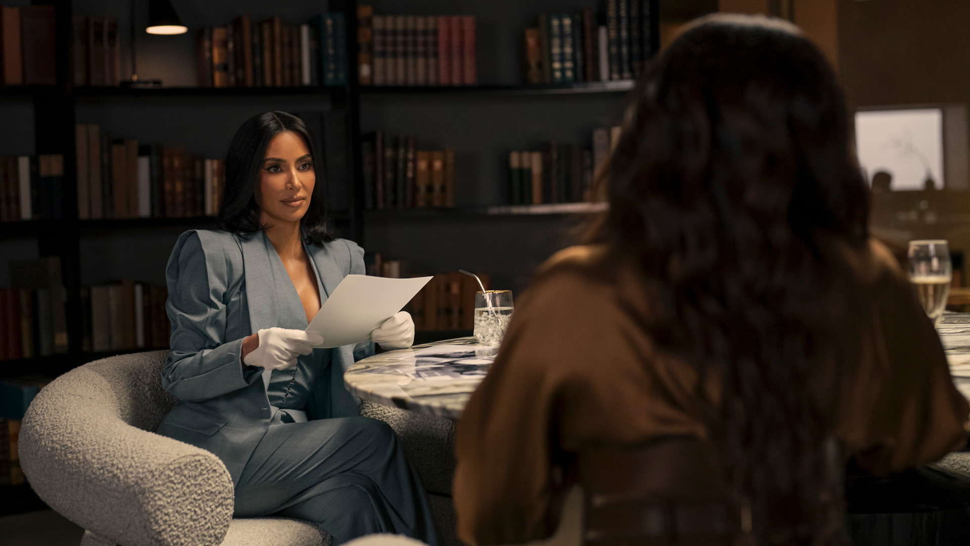 In dieser Szene aus All's Fair sitzt Kim Kardashian als Allura Grant in einem eleganten grauen Anzug mit weißen Handschuhen an einem runden Tisch. Sie hält konzentriert ein Dokument in den Händen und wirkt ernst und fokussiert. Im Vordergrund ist unscharf eine weitere Person zu sehen. Die Stimmung ist angespannt und geschäftlich.