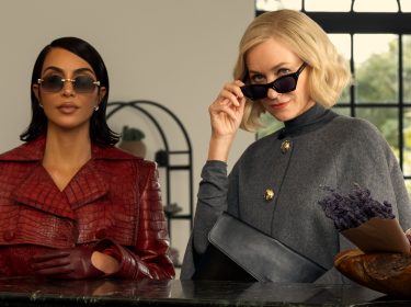 In dieser stilvollen Szene aus All's Fair stehen Kim Kardashian als Allura Grant und Naomi Watts als Liberty Ronson nebeneinander. Allura trägt einen roten Mantel in Krokodiloptik mit passenden Handschuhen und Sonnenbrille, während Liberty in einem grauen Cape und mit hochgezogener Sonnenbrille selbstbewusst in die Kamera blickt. Die Stimmung ist kühl, kontrolliert und voller unterschwelliger Spannung.