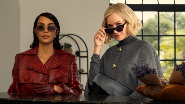 In dieser stilvollen Szene aus All's Fair stehen Kim Kardashian als Allura Grant und Naomi Watts als Liberty Ronson nebeneinander. Allura trägt einen roten Mantel in Krokodiloptik mit passenden Handschuhen und Sonnenbrille, während Liberty in einem grauen Cape und mit hochgezogener Sonnenbrille selbstbewusst in die Kamera blickt. Die Stimmung ist kühl, kontrolliert und voller unterschwelliger Spannung.