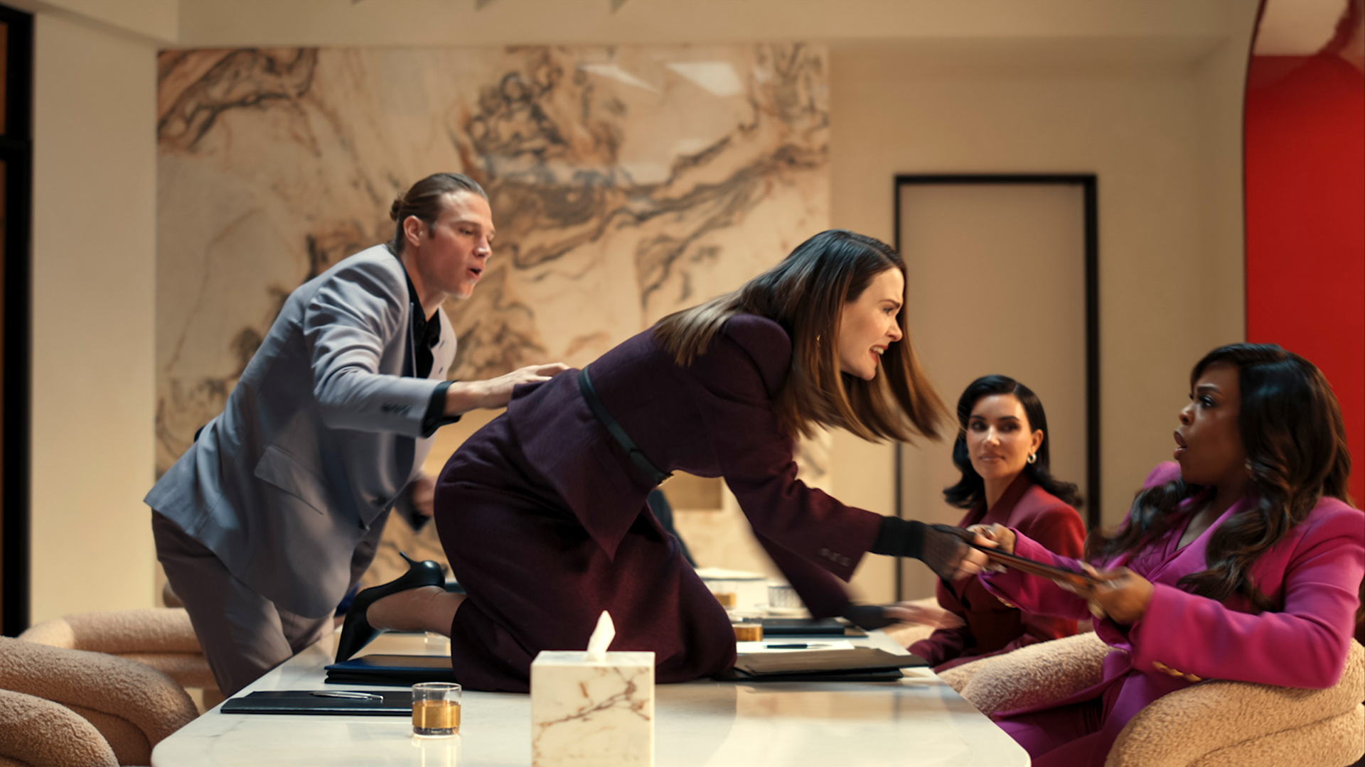 In dieser dramatischen Szene aus All's Fair kniet Sarah Paulson als Carr Lane wütend auf einem Konferenztisch und streitet mit Niecy Nash-Betts als Emerald Greene, die erschrocken zurückweicht. Links versucht Alluras Exmann, die Situation zu beruhigen, während Kim Kardashian als Allura Grant das Geschehen angespannt beobachtet. Die Stimmung ist explosiv und voller Spannung.