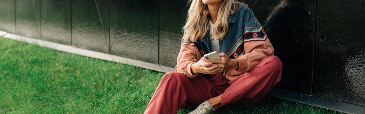 Frau sitzt auf Hügel mit Handy in der Hand und AirPods in den Ohren
