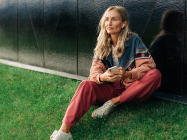 Frau sitzt auf Hügel mit Handy in der Hand und AirPods in den Ohren