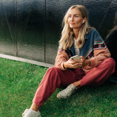 Frau sitzt auf Hügel mit Handy in der Hand und AirPods in den Ohren