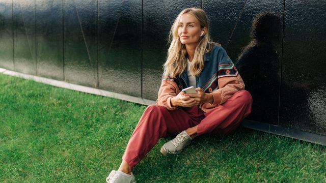 Frau sitzt auf Hügel mit Handy in der Hand und AirPods in den Ohren