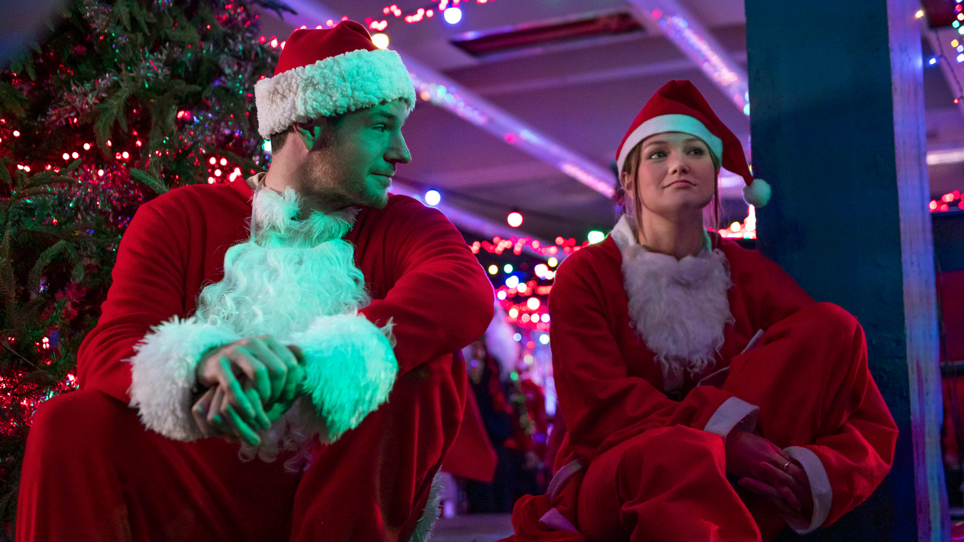 Im Bild aus Jingle Bell Heist sitzen Olivia Holt als Sophia und Connor Swindells als Nick nebeneinander in Weihnachtsmannkostümen. Die beiden wirken erschöpft, aber verbunden, während sie sich inmitten von bunten Lichterketten und einem geschmückten Weihnachtsbaum anschauen. Die Szene aus dem Netflix-Film Jingle Bell Heist kombiniert festliche Atmosphäre mit emotionaler Tiefe.