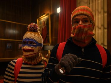 Im Bild aus Jingle Bell Heist tragen Olivia Holt als Sophia und Connor Swindells als Nick skurrile, selbstgestrickte Sturmhauben mit bunten Mustern. In gestreiften Pullovern, roten Rucksäcken und mit Taschenlampe bewaffnet, wirken sie wie improvisierte Einbrecher. Die Szene aus dem Netflix-Weihnachtsfilm Jingle Bell Heist kombiniert Spannung mit einem humorvollen Ton.