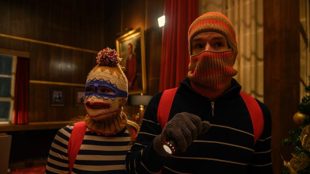 Im Bild aus Jingle Bell Heist tragen Olivia Holt als Sophia und Connor Swindells als Nick skurrile, selbstgestrickte Sturmhauben mit bunten Mustern. In gestreiften Pullovern, roten Rucksäcken und mit Taschenlampe bewaffnet, wirken sie wie improvisierte Einbrecher. Die Szene aus dem Netflix-Weihnachtsfilm Jingle Bell Heist kombiniert Spannung mit einem humorvollen Ton.