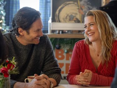 A Merry Little Ex-Mas: Ein Mann und eine Frau unterhalten sich lächelnd an einem Tisch in einem weihnachtlich geschmücktem Wohnzimmer.