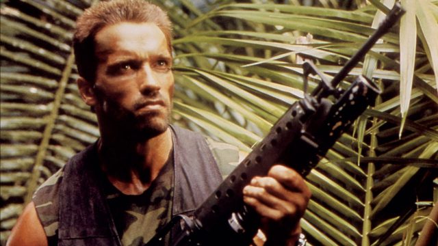 Arnold Schwarzenegger als Alan „Dutch“ Schaefer im Film "Predator". Er steht im Dschungel vor einer Pflanze und hält ein Schnellfeuergewehr in der Hand.